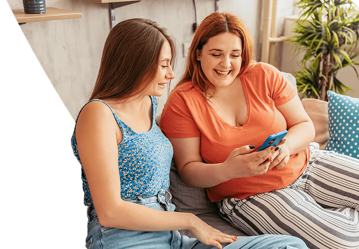 Two young women are seated on a sofa in a living room, with one of them displaying her phone to the other, and both are smiling.