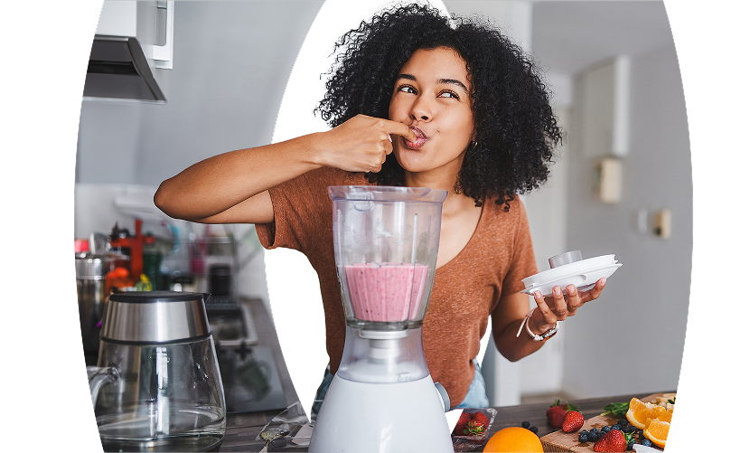Young curly-haired women in her kitchen, smiling as she tastes a smoothie with her finger, with a juicer machine and fresh fruits on the table