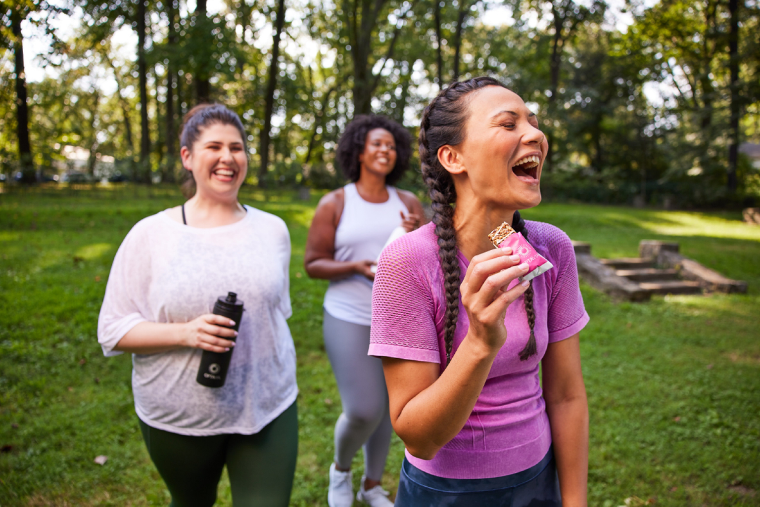 Three woman walking outdoors smiling. Woman in front is holding an Optavia Fueling bar.